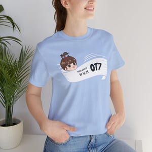 Pode incluir: T-shirt azul claro com um desenho animado de uma menina e o texto "OT7 with extra W.W.H." A t-shirt de manga curta é feita de tecido macio. O design sugere que está relacionado com merchandising de fãs ou um grupo específico.