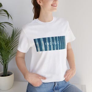 May include: White t-shirt with a rectangular design featuring vertical blue stripes and the word "INDIGO" in white. The shirt is short-sleeved and made of a soft material.