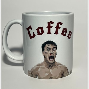 May include: White ceramic coffee mug with a handle. The mug features a graphic of a man with a bloody nose and an open mouth, with the word "Coffee" in maroon above the image. The mug is a standard size.