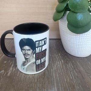 May include: White ceramic mug with a black handle and interior. The mug features a portrait of a man with a mustache and the text "IT AIN'T MY FIRST OR MY LAST". A plant in a white pot sits to the right.