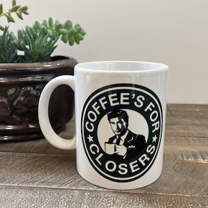 May include: White ceramic coffee mug with a green circle design featuring a black silhouette of a man in a suit holding a coffee cup. The text "Coffee's For Closers" is written in black around the circle.