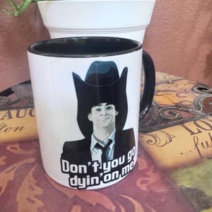May include: White ceramic mug with a black interior and handle. The mug features a graphic of a person wearing a cowboy hat and suit, with the text "Don't you go dyin' on me."