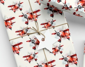 May include: Wrapping paper featuring a repeating watercolor pattern of red biplane aircraft on a cream background. The paper is tied with brown twine and includes white star and heart-shaped gift tags, a ball of twine, and black scissors.