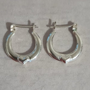 May include: A pair of polished silver hoop earrings featuring a small, raised heart detail at the base of each hoop. The earrings are designed with a hinged post closure mechanism, providing a secure fit for the wearer while maintaining a smooth finish.