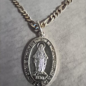 May include: Silver-toned necklace with a detailed oval pendant. The pendant features an engraved image of a religious figure and the text "O Mary Conceived Without Sin Pray For Us Who Have Recourse To Thee 1830". The chain has a textured design.