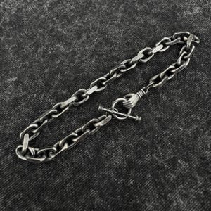 Peut inclure: Bracelet en chaîne argentée avec fermoir à bascule. Le bracelet présente une série de maillons ovales interconnectés. Le fermoir est une main tenant une barre. Le bracelet est présenté sur une surface texturée gris foncé.