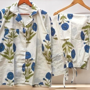 Könnte beinhalten: Weißes Pyjama-Set mit blauem Blumenmuster und grünen Blättern. Das Set besteht aus einem Hemd mit Knöpfen und einer Hose mit Kordelzug. Die Pyjamas hängen an Holzkleiderbügeln.