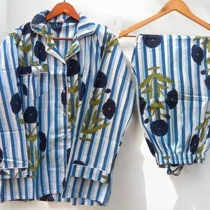 May include: Blue and white striped pyjama set with a floral pattern. The set includes a button-down shirt with a chest pocket and long trousers. The floral design features navy blue flowers and green leaves. The pyjamas are hanging on wooden hangers.