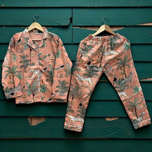 Könnte beinhalten: Pfirsichfarbenes Pyjama-Set mit Dschungel-Motiv. Das Set besteht aus einem Hemd mit Knöpfen und einer Hose, beide mit Zebras, Leoparden, Vögeln und Palmen verziert. Die Pyjamas werden auf Holzbügeln vor einem grünen Hintergrund präsentiert.