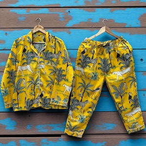 May include: A yellow pajama set with a jungle print featuring zebras, leopards, toucans, and palm trees. The button-down shirt and drawstring trousers are displayed on wooden hangers against a blue and brown background.