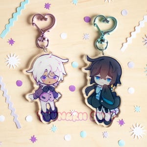 Puede incluir: Dos llaveros acrílicos estilo anime con cierres en forma de corazón. Un llavero presenta un personaje con cabello blanco y un atuendo morado, el otro tiene un personaje con cabello oscuro y un atuendo negro. El fondo es de madera clara con confeti.