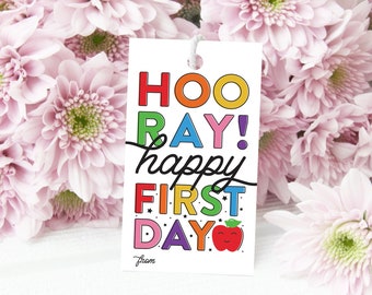 Könnte beinhalten: Ein weißer Geschenkanhänger mit dem Text "HOORAY! happy FIRST DAY" in Regenbogenfarben mit einem roten Apfel mit einem lächelnden Gesicht. Der Text "from" befindet sich am unteren Rand des Anhängers.
