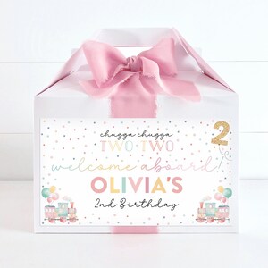 Puede incluir: Una caja de regalo blanca con una cinta rosa y una etiqueta colorida que dice "chugga chugga TWO TWO welcome aboard! OLIVIA'S 2nd Birthday".
