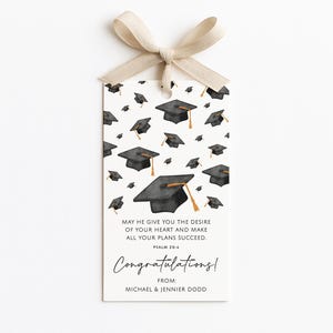 Op de afbeelding: Een afstudeergeschenktag met een beige lint. De tag heeft een patroon van zwarte afstudeerpetten met gouden kwastjes op een witte achtergrond. Tekst bevat "May He give you the desire of your heart..." en "Congratulations!"