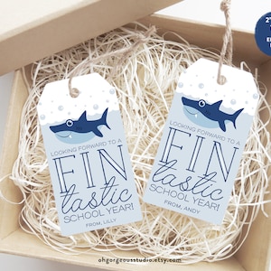 May include: Two blue and white shark-themed gift tags with the text "Looking forward to a Fin-tastic School Year!" and the names "Lilly" and "Andy". The tags are 2 inches by 3.5 inches and are editable.