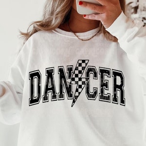 Könnte beinhalten: Weißer Pullover mit schwarzem Text, der "DANCER" mit einem karierten Blitz-Motiv sagt.