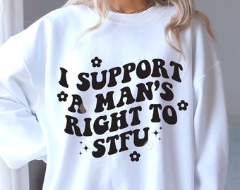 Op de afbeelding: Witte trui met zwarte tekst die "I support a man's right to STFU" luidt met kleine bloemen- en sterretjes.