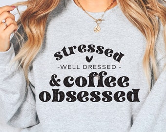 May include: A light gray sweatshirt with the words "stressed - well dressed - & coffee obsessed" in black lettering. The sweatshirt has a crew neck and long sleeves. The text is in a mix of fonts.