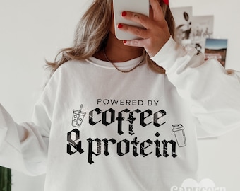 May include: White sweatshirt with black text that reads "Powered by coffee & protein". The text is in a gothic font and includes a graphic of a coffee cup and a protein shaker.
