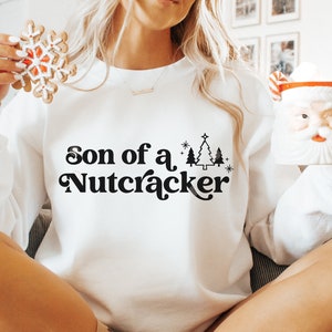 May include: White sweatshirt with the text "Son of a Nutcracker" in black script. The sweatshirt also features a Christmas tree graphic. The person is holding a snowflake cookie and a Santa mug.