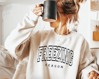 May include: A cream-colored sweatshirt with the text "FREEZING SEASON" in black block letters. The sweatshirt is worn by someone holding a black mug. The image also includes the text "PNG + SVG".