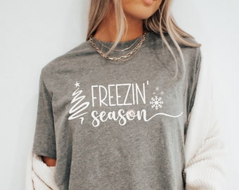 May include: A gray t-shirt with the text "Freezin' Season" in white, featuring a stylized Christmas tree and snowflake. The shirt is a casual style, suitable for winter or holiday wear.