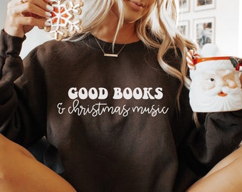 May include: A woman wearing a brown sweatshirt with the text "GOOD BOOKS & christmas music" in white. She is sitting on a white fuzzy blanket and holding a white mug with a Santa Claus design.