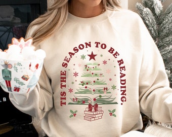 May include: A cream-coloured sweatshirt with the text "TIS THE SEASON TO BE READING." The design features a Christmas tree made of books, with a star on top. The sweatshirt has a drawstring at the waist.