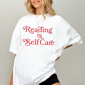Puede incluir: Camiseta blanca de gran tama&ntilde;o con el texto "Reading is Self Care" en letras rojas.