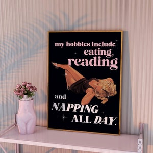 May include: A framed black poster with gold trim displays the text "my hobbies include eating, reading, and napping all day." The poster has a vintage illustration of a woman. A pink vase with flowers sits on a pink shelf.