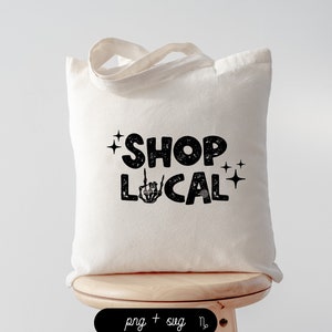 Op de afbeelding: Een crèmekleurige draagtas met de tekst "SHOP LOCAL" in een vetgedrukte, zwarte letter. De letter "O" in "SHOP" is een ster en de letter "O" in "LOCAL" is een skelet hand die de middelvinger opsteekt. De tas heeft een eenvoudig ontwerp met twee handvatten.