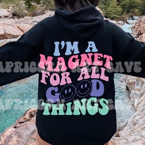 May include: A black hooded sweatshirt with the text "I'm a magnet for all good things" in pink, blue, and green lettering with smiley faces. The person wearing the sweatshirt is standing in front of a river and rocks.