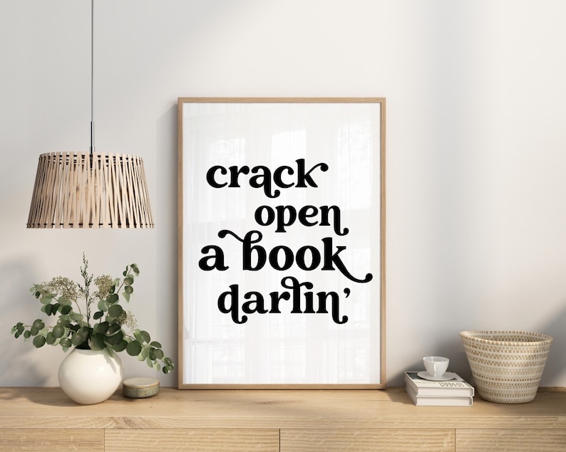 May include: A black and white typography print with the text "crack open a book darlin'" in a retro font style. The print is framed in a light wood frame and is hanging on a white wall.