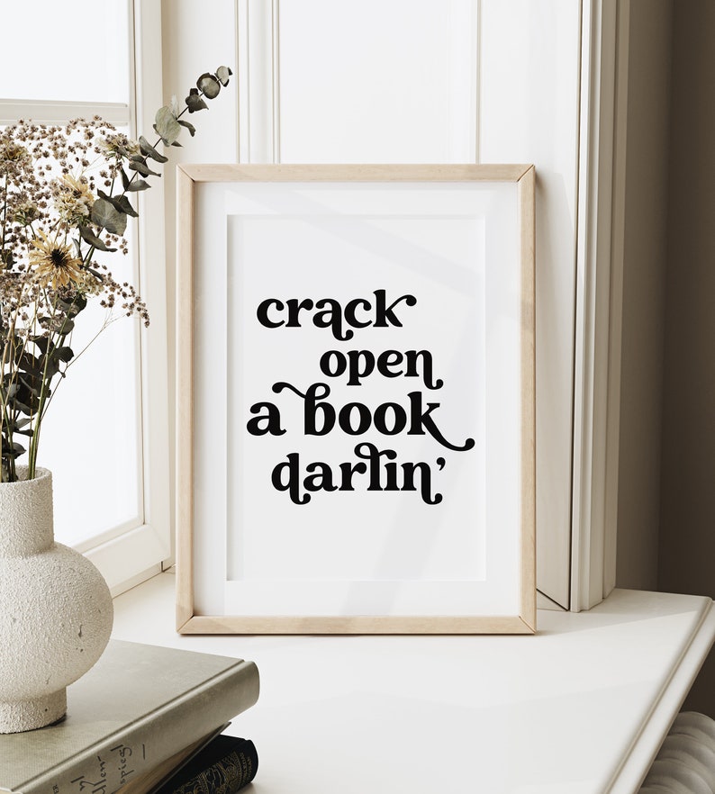 May include: A framed print with a white background and black text that reads "crack open a book darlin'" in a retro font style.