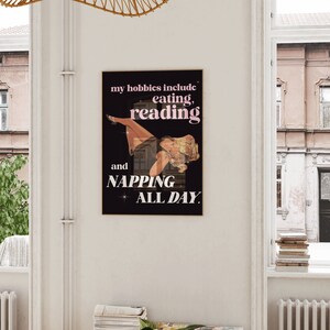 May include: A framed poster with a black background and the text "my hobbies include eating, reading, and napping all day." The image features a vintage illustration of a woman lounging in a window. The text is in white and pink.
