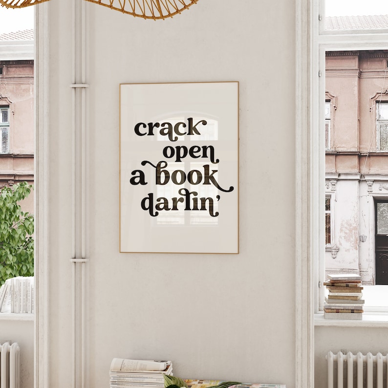 May include: A white framed print with black text that reads "crack open a book darlin'" hanging on a white wall.