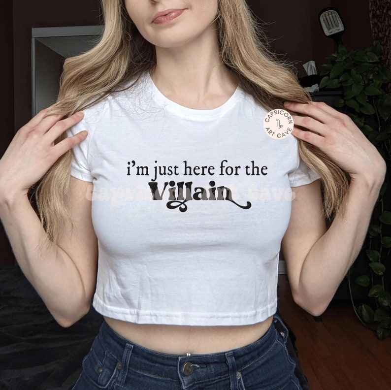 May include: White crop top with the text "I'm just here for the Villain" in black and gray. The shirt is a crew neck style and is paired with blue jeans. A small logo is visible in the upper right corner.