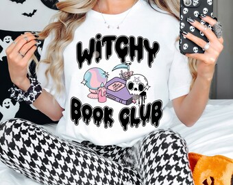 Peut inclure: T-shirt blanc avec l'inscription "Witchy Book Club" en noir, dans une police de caractères arrondie. Le t-shirt présente un dessin d'une boule de cristal, d'un livre, d'un crâne et d'une bougie. Le design a des couleurs pastel et un thème effrayant.