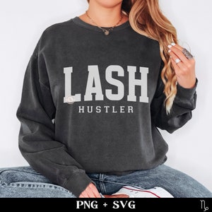 May include: Dark grey sweatshirt featuring the white text "LASH HUSTLER". The text is in a bold, block font. The sweatshirt has a faded, vintage appearance. The words are centered on the chest.