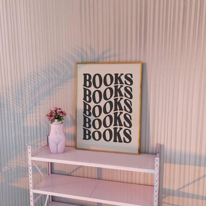 May include: A gold framed print with the word "BOOKS" repeated five times in a black retro font. The print is on a white shelf with a pink vase with pink flowers on the shelf.