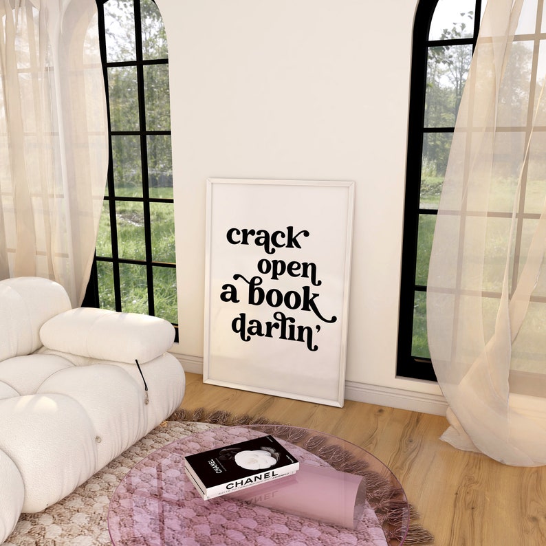 May include: A white framed print with black text that reads "crack open a book darlin'" hanging on a white wall in a room with a white couch and a glass coffee table with a book on it.