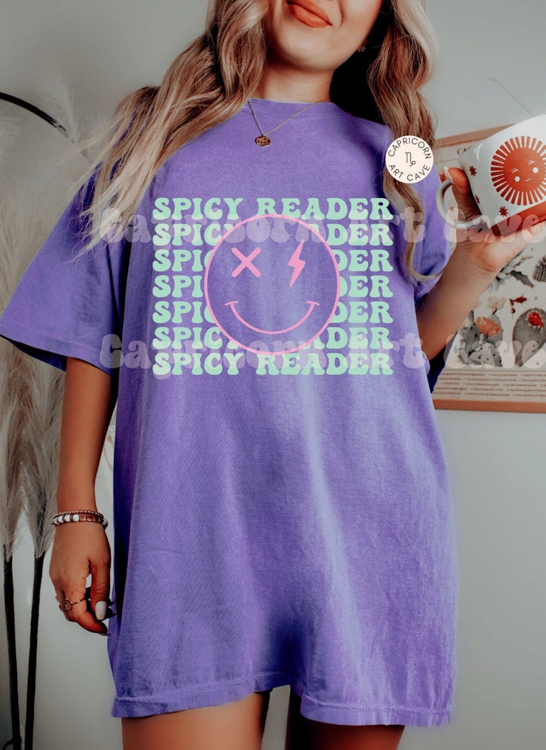 May include: A purple t-shirt with the text "SPICY READER" in a stacked design. A smiling face graphic with an X for eyes and a lightning bolt is in the center. The shirt is a solid color and has a relaxed fit.
