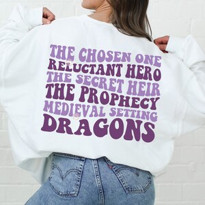 以下が含まれることがあります： 白いスウェットシャツに、紫色の文字で「The Chosen One, Reluctant Hero, The Secret Heir, The Prophecy, Medieval Setting, Dragons」と書かれています。