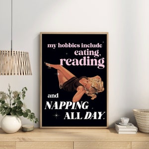 May include: A framed poster with a black background and a vintage illustration of a woman reclining. The text reads "my hobbies include eating, reading, and napping all day."