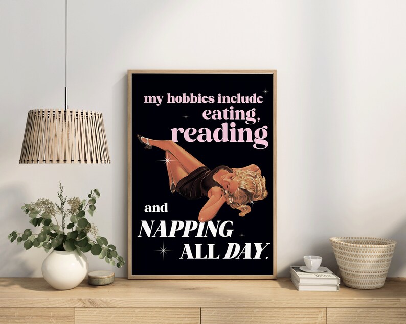 May include: A framed poster with a black background and a vintage illustration of a woman reclining. The text reads "my hobbies include eating, reading, and napping all day."