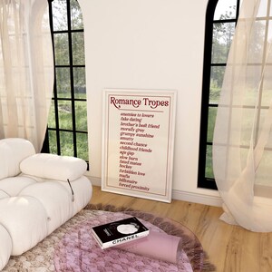 Puede incluir: Un p&oacute;ster enmarcado blanco con el t&iacute;tulo "Romance Tropes" y una lista de tropos rom&aacute;nticos comunes en texto rojo. El p&oacute;ster est&aacute; colgado en una pared blanca en una habitaci&oacute;n con un sof&aacute; blanco y una mesa de vidrio.