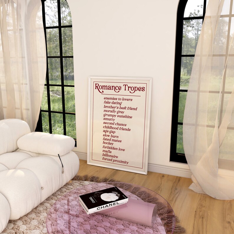 Puede incluir: Un p&oacute;ster enmarcado blanco con el t&iacute;tulo "Romance Tropes" y una lista de tropos rom&aacute;nticos comunes en texto rojo. El p&oacute;ster est&aacute; colgado en una pared blanca en una habitaci&oacute;n con un sof&aacute; blanco y una mesa de vidrio.