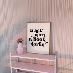May include: A white framed print with black text that reads "crack open a book darlin'" on a white shelf with a pink ceramic vase with pink flowers.