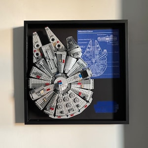 May include: A framed LEGO Millennium Falcon model with a blueprint of the ship in the background. The model is made of gray and blue LEGO bricks and features a detailed interior. The frame is black and has a white mat.