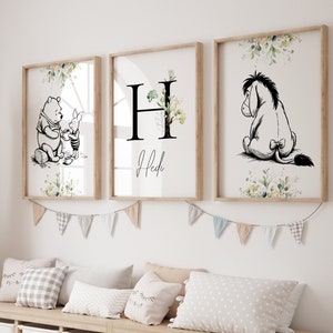 May include: Three framed prints with a Winnie the Pooh theme. The first print features Winnie the Pooh and Piglet, the second print features the letter H with floral accents and the name "Heidi" below, and the third print features Eeyore. The prints are hanging on a white wall above a wooden shelf with storage bins.